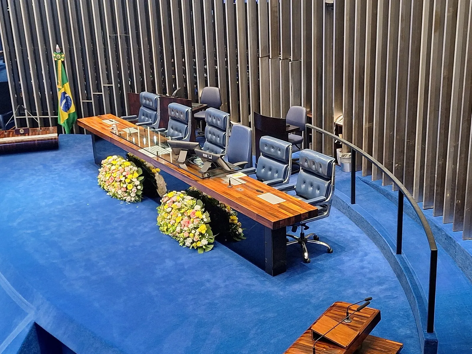 Mesa diretora vazia do Plenário do Senado Federal, com cadeiras azuis, bandeira do Brasil e coroas de flores.