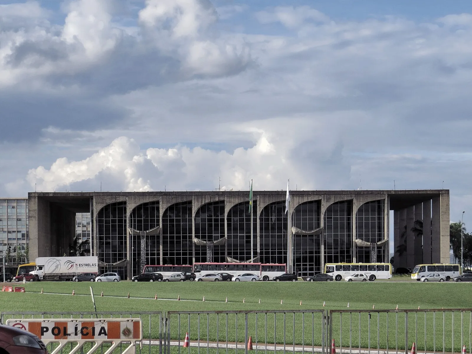 Fachada de concreto e vidro, com grandes arcos, do Palácio da Justiça em Brasília, com ônibus e carros em frente e uma placa de 'Polícia' na cerca em primeiro plano.