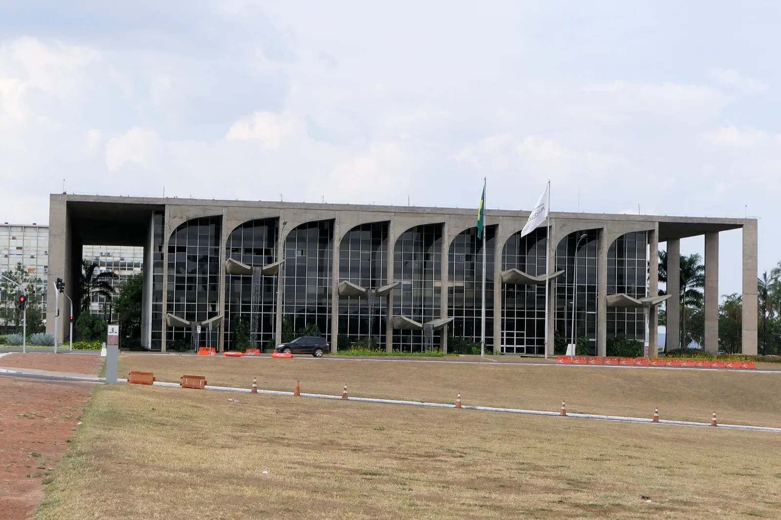 Fachada de prédio de concreto com arcos e janelas de vidro em Brasília, com bandeira do Brasil e bandeira do Mercosul no mastro, gramado em primeiro plano.