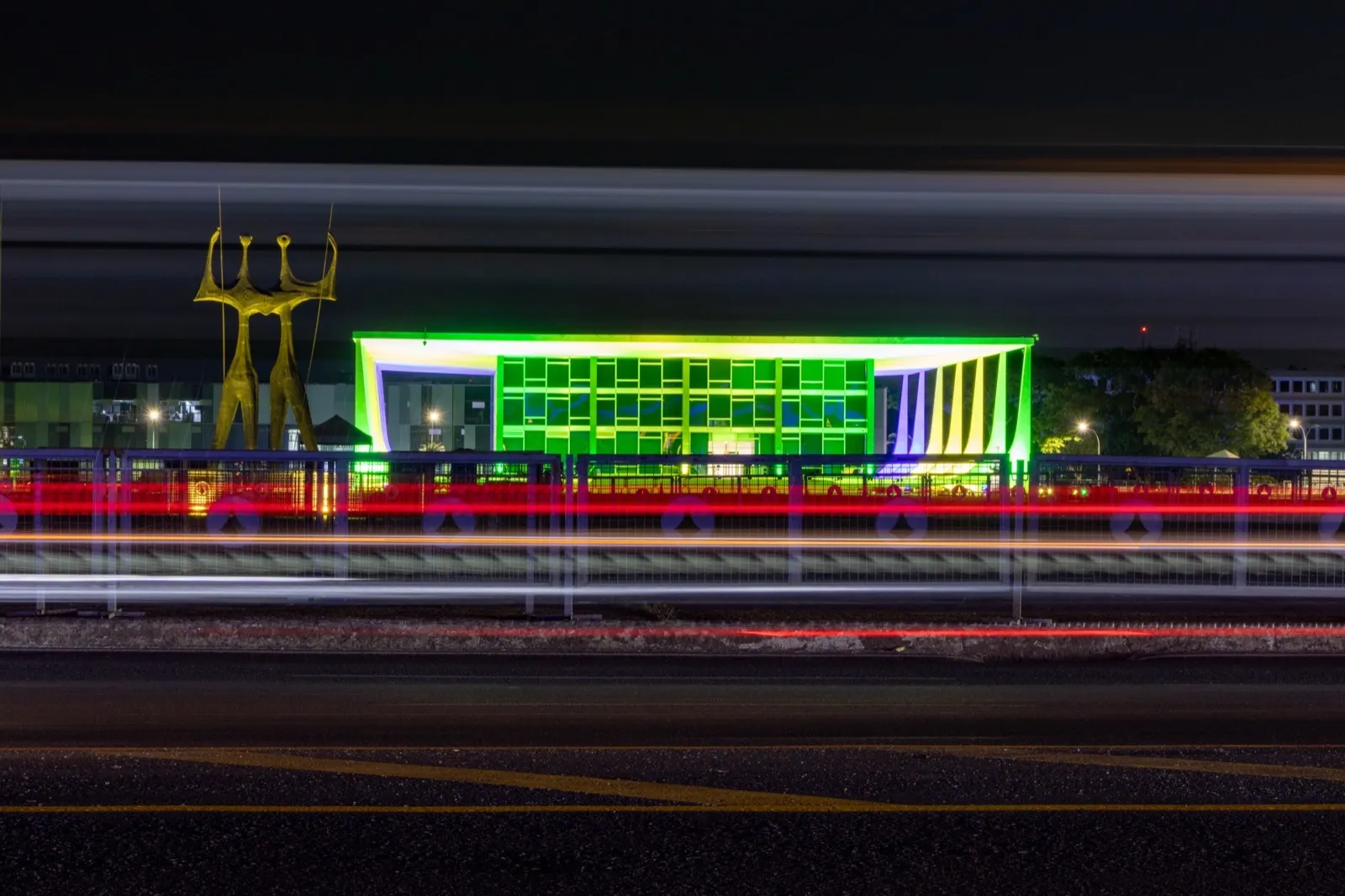 Fachada do Supremo Tribunal Federal iluminada em verde e amarelo, à noite, em Brasília, com a escultura Os Candangos à esquerda.