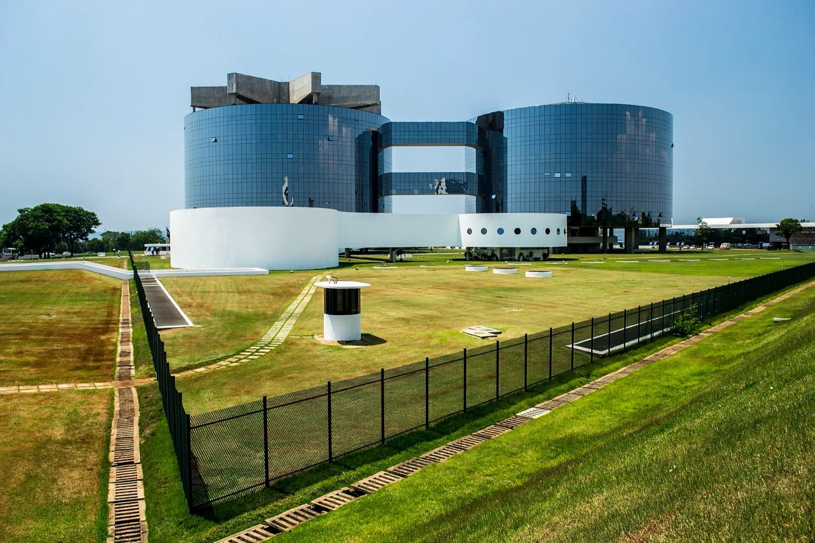 Fachada da sede da Procuradoria-Geral da República, em Brasília, com dois grandes cilindros de vidro azul, um bloco branco curvo e gramado com cerca preta em primeiro plano.