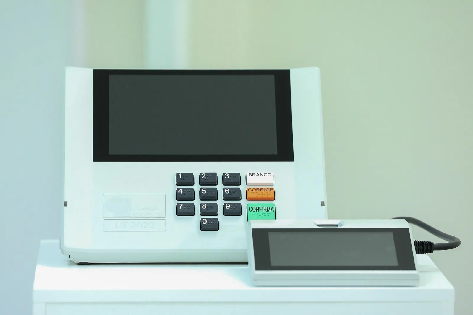 Urna eletrônica modelo UE2020 sobre uma bancada branca, com teclado numérico e as teclas BRANCO, CORRIGE e CONFIRMA, com a tela apagada.