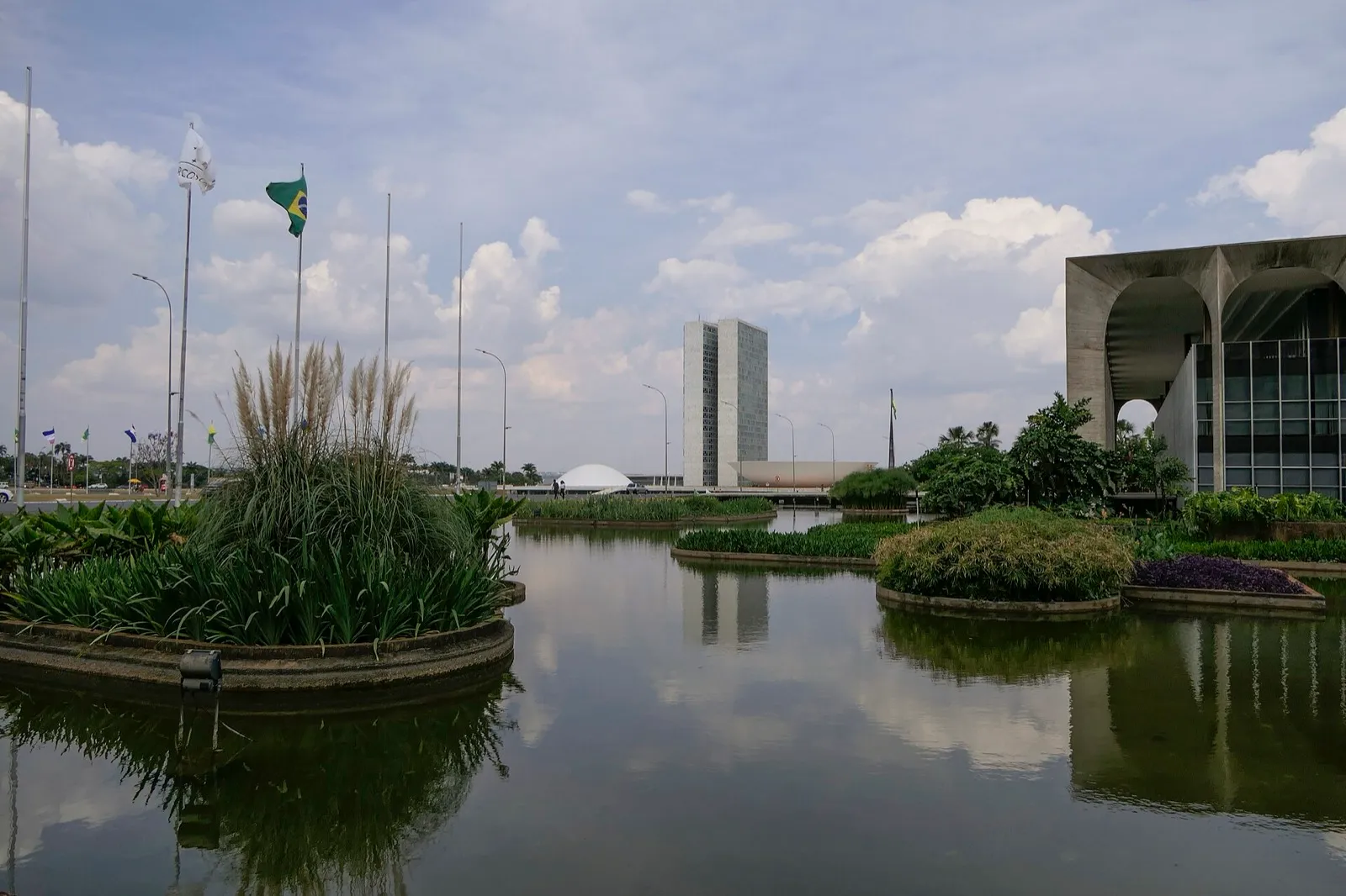 Vista do Congresso Nacional, em Brasília, com as duas torres e a cúpula ao fundo, sobre um espelho d'água, e um prédio de arcos à direita.