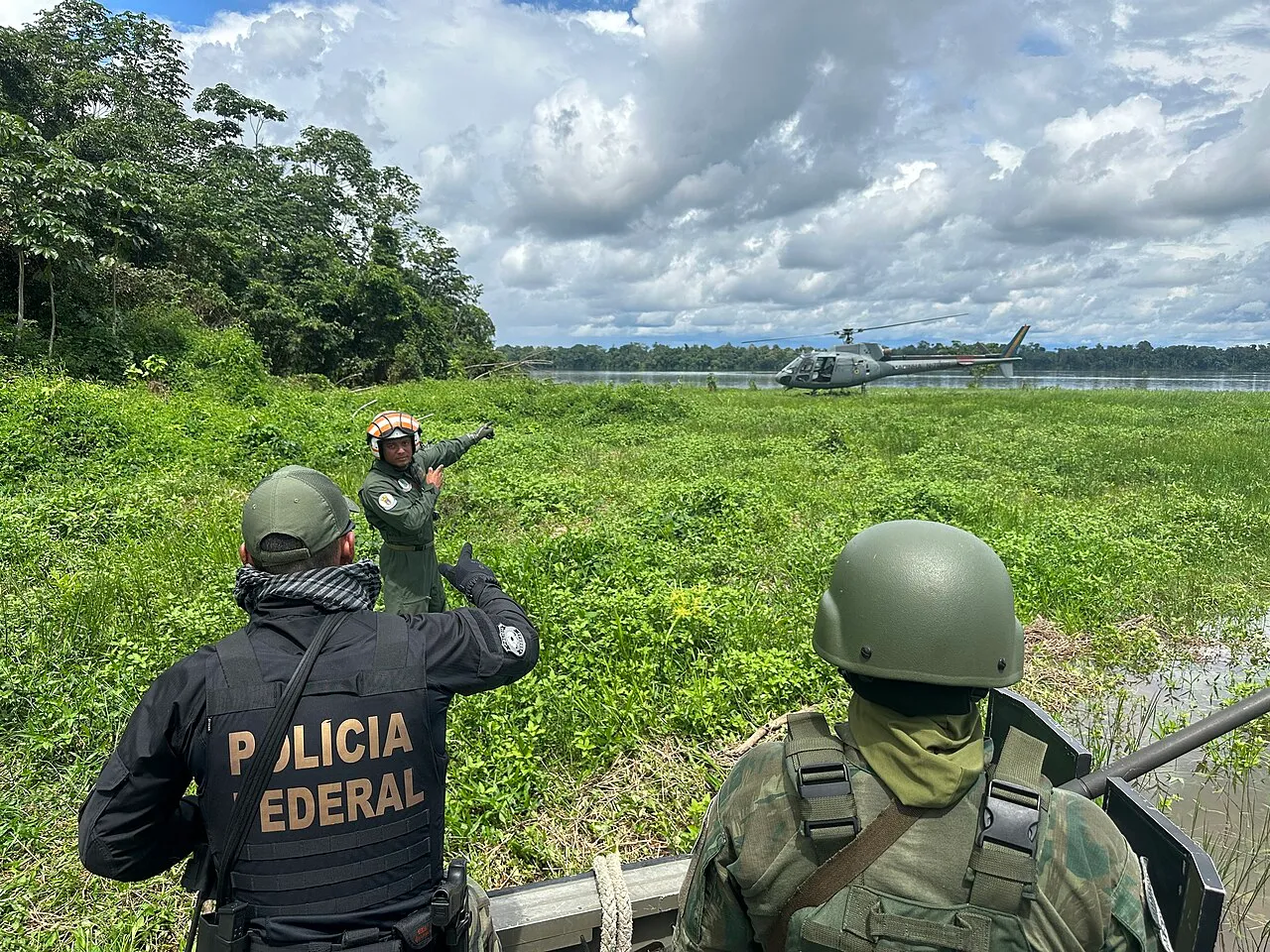 Agente da Polícia Federal de colete com a sigla da corporação e militar de capacete observam um helicóptero ao fundo, perto de um rio na Amazônia.