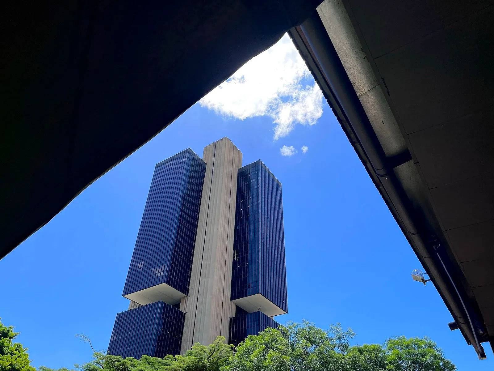 Torres de vidro escuro e um pilar de concreto do prédio-sede do Banco Central, em Brasília, vistos de baixo contra o céu azul.
