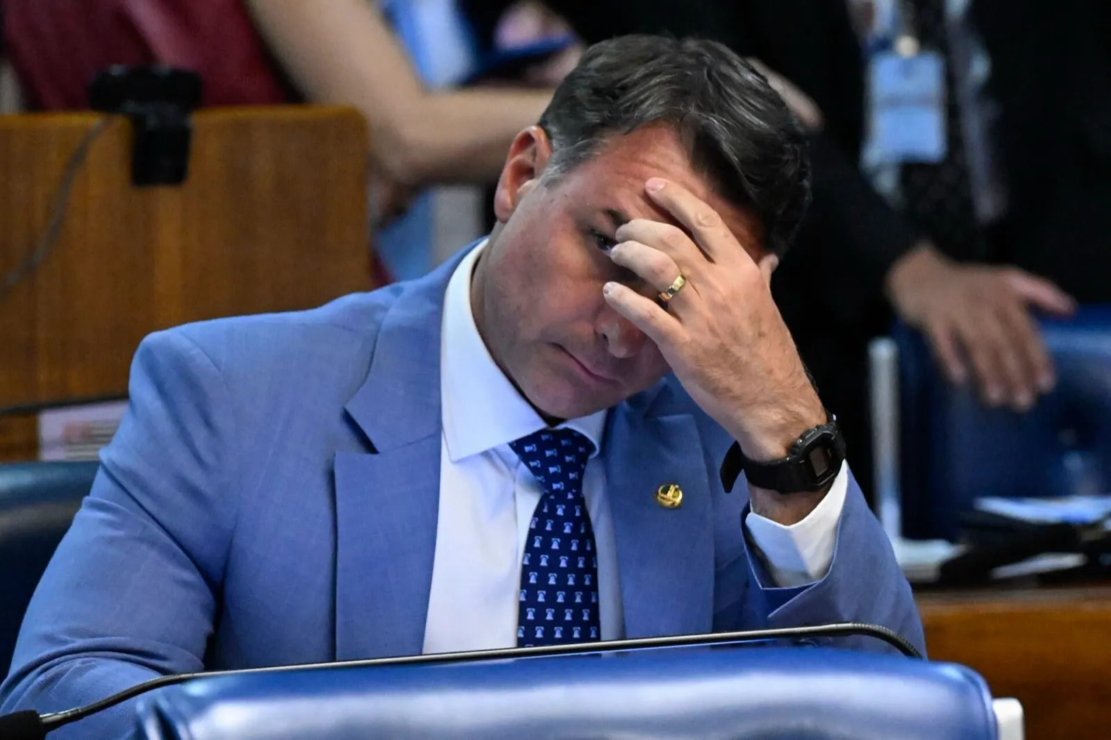 Flávio Bolsonaro, de terno azul-claro e gravata azul, sentado à bancada do Plenário do Senado, com a mão na testa.