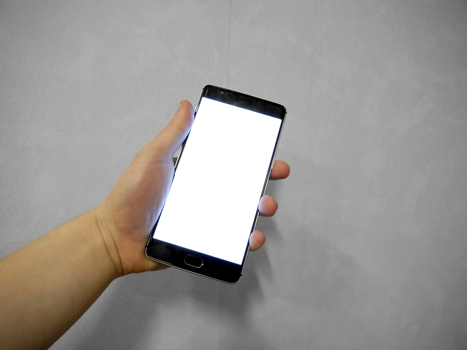 Mão de uma pessoa segurando um smartphone com tela branca em branco, contra um fundo cinza liso.