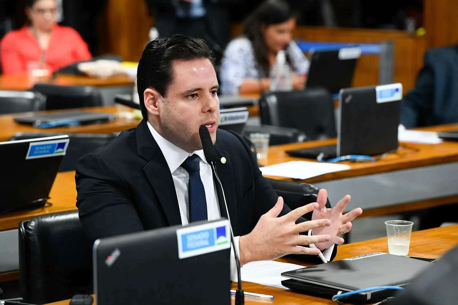 O deputado Rodrigo Valadares, de terno escuro e gravata azul, fala ao microfone sentado à bancada de uma comissão do Congresso, gesticulando com as mãos.