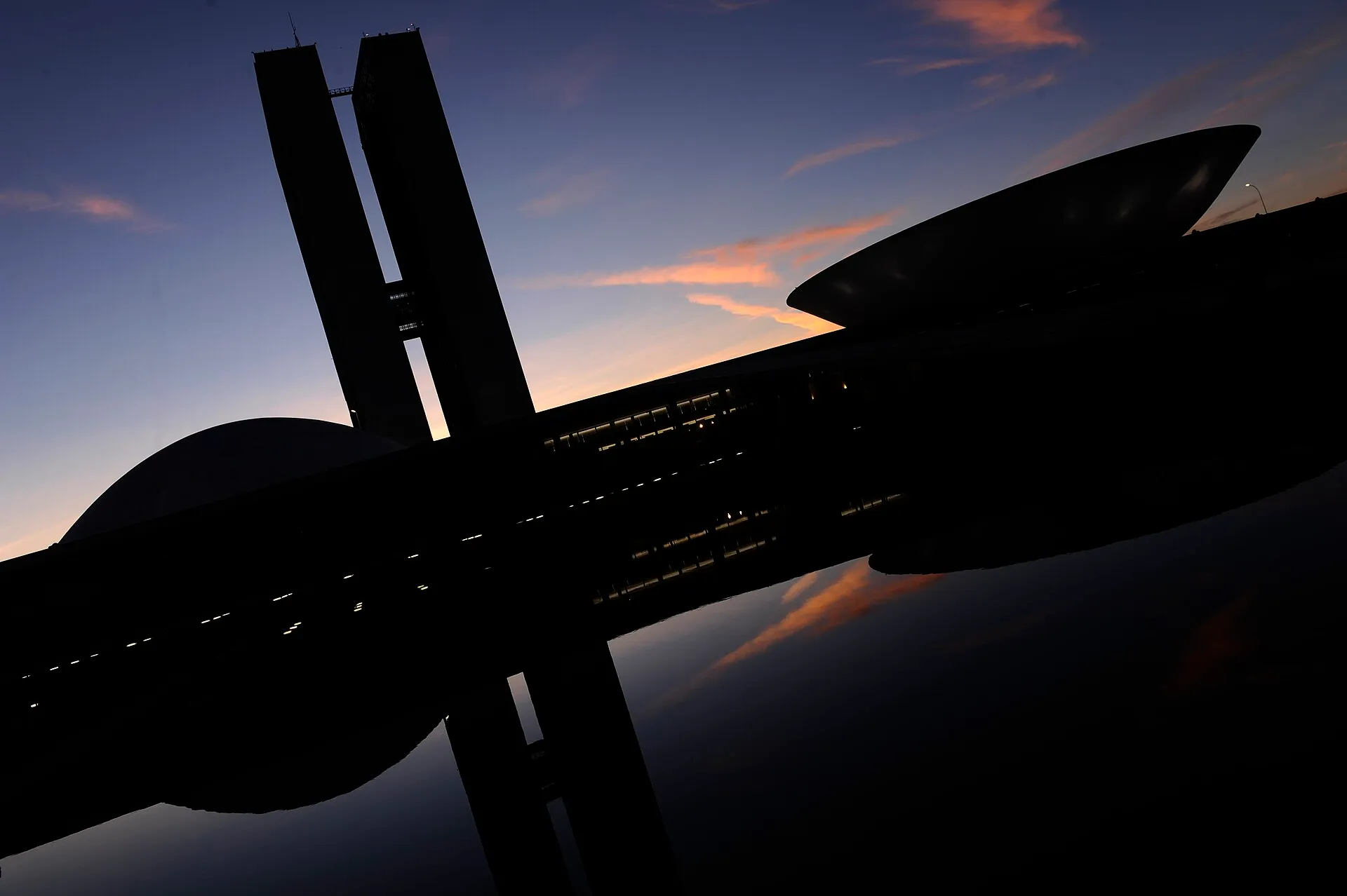 Fachada do Congresso Nacional, em Brasília, em silhueta contra o céu, com reflexo na água em primeiro plano.