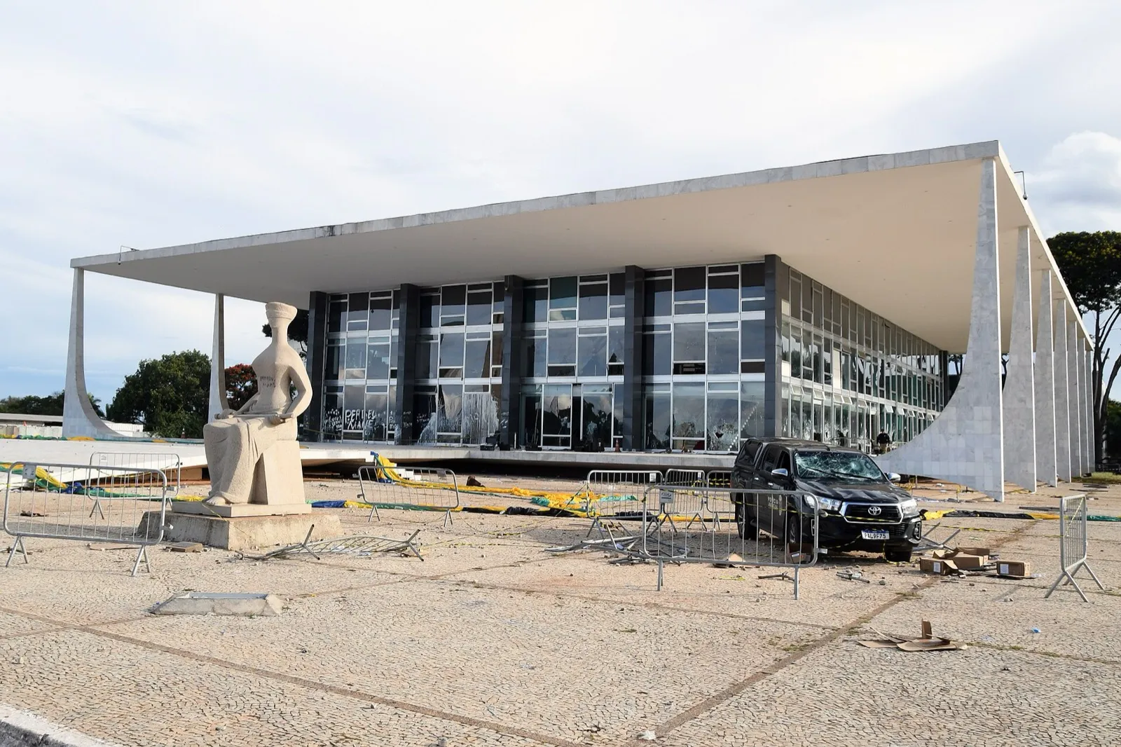 Fachada do prédio do STF com vidros quebrados e pichações, grades derrubadas na praça e uma caminhonete com o para-brisa estilhaçado; à esquerda, a estátua da Justiça pichada.
