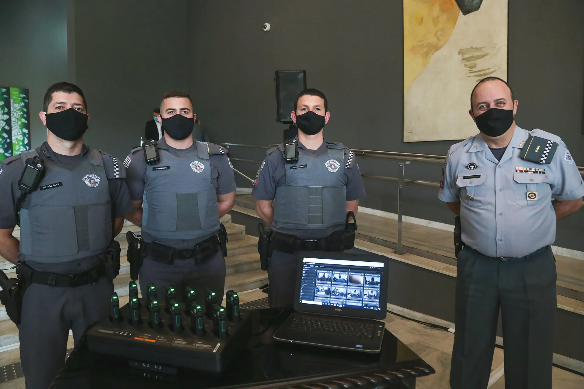 Três soldados e um oficial da Polícia Militar de São Paulo, de máscara preta e farda, com câmeras corporais presas ao uniforme, ao lado de carregadores de câmeras e de um notebook com imagens de gravações.