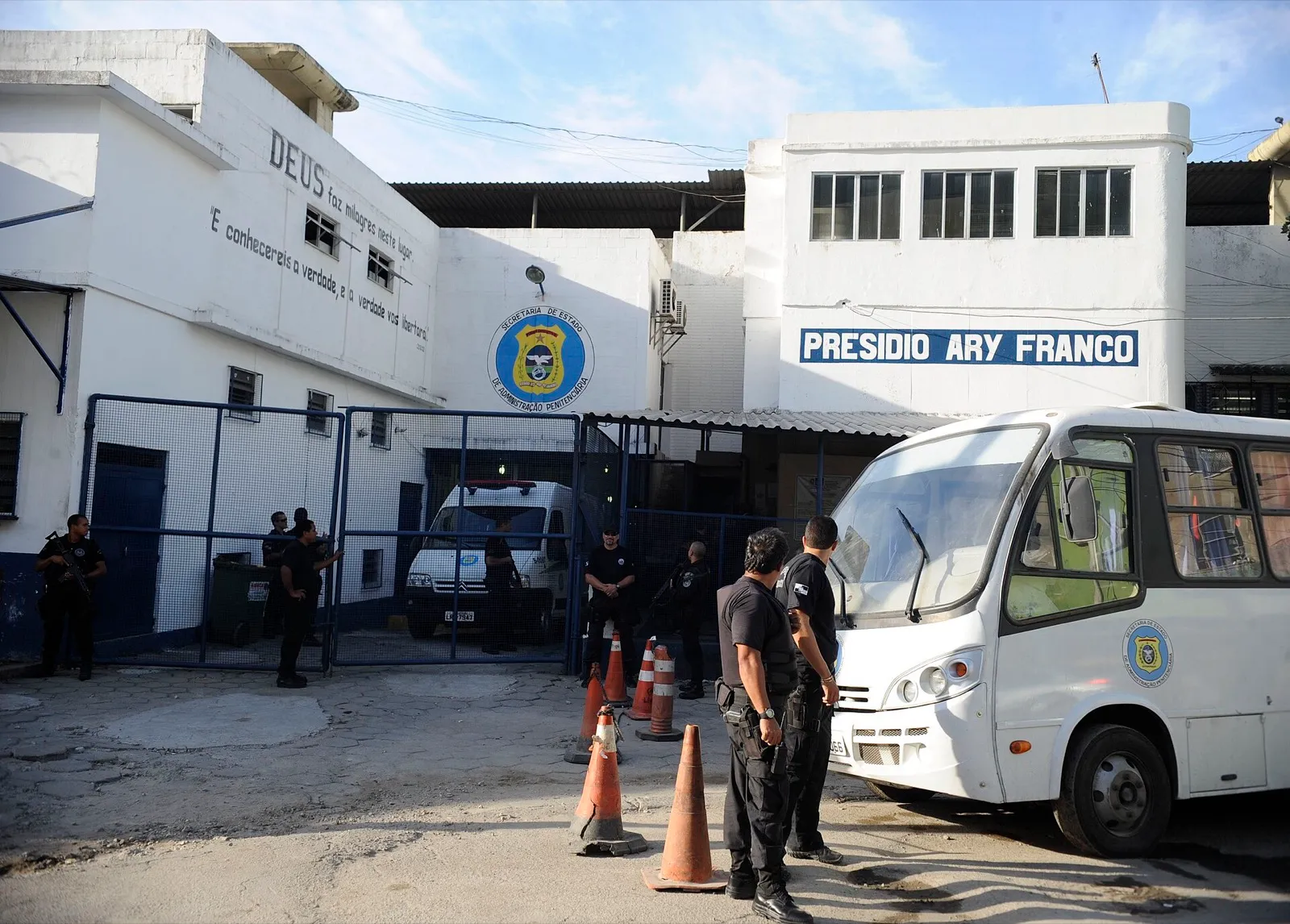 Fachada do Presídio Ary Franco, no Rio de Janeiro, com a placa da Secretaria de Administração Penitenciária, agentes armados no portão e um ônibus branco de transporte de presos.
