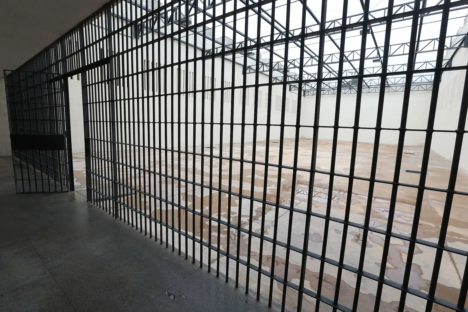 Grade de ferro de um pátio de banho de sol vazio, com muros altos e cobertura de tela metálica, em presídio de segurança máxima.